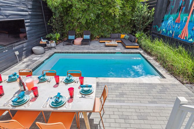 Modern backyard patio with rectangular swimming pool, outdoor dining table set with teal plates and red cups, orange chairs, lounge seating, bamboo privacy screen and colorful mural