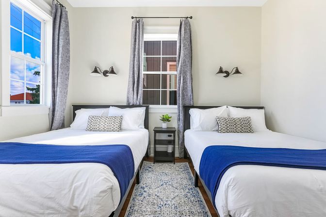 Bright twin bedroom with two white beds dressed in navy throws and patterned pillows, gray curtains framing a central window, black bedside table with potted plant, wall sconces and light hardwood floor.