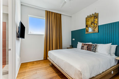 Sunlit, chic modern bedroom with a king-size bed in white linens, teal beadboard headboard, geometric accent pillow, mustard curtains by a small window, wall-mounted TV, hardwood floors and framed musician artwork.
