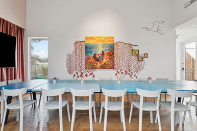 Bright beachside meeting room with long turquoise tables and white chairs, colorful ocean sunset painting framed by red coral-pattern wall art, wood floors, seagull line drawing and sunlit windows.