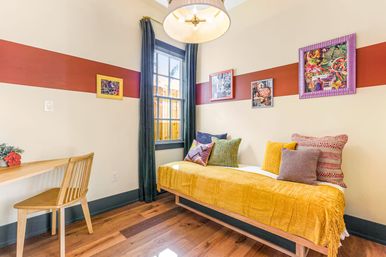 Sunlit cozy guest room with a mustard-yellow daybed topped with colorful textured pillows, warm wood floors, a small wooden desk and chair, a dark-trim window with curtains, and a red horizontal accent stripe with framed artwork on pale walls.