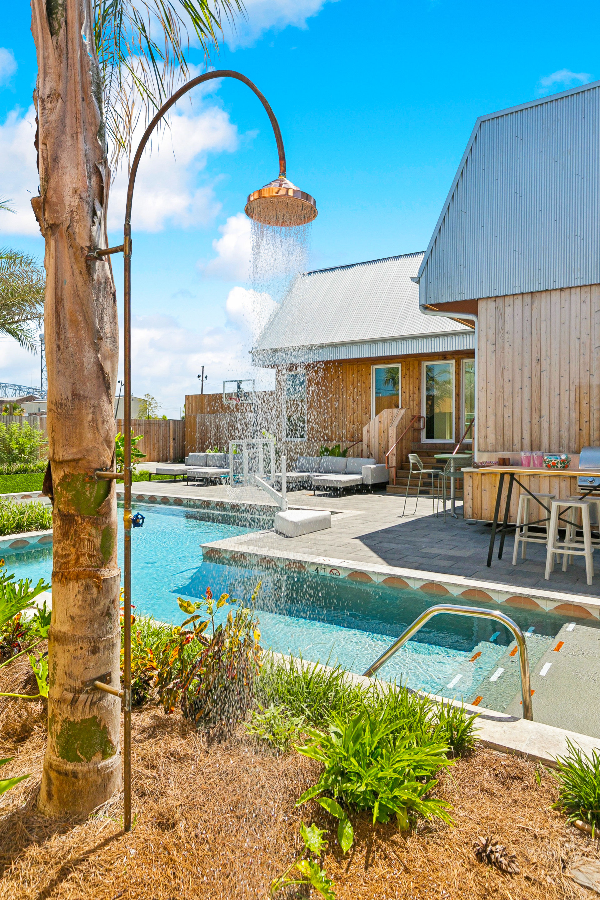 Copper outdoor shower dumping water into a turquoise backyard pool beside a modern wooden pool house, lounge seating, bar stools and tropical landscaping on a sunny day