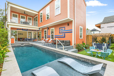 Sunny modern backyard with rectangular pool and submerged lounge chairs, orange two-story house with balcony and covered patio, outdoor dining and lounge seating, blue geometric wall mural and tropical landscaping.