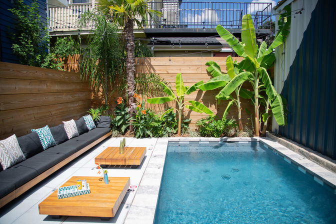 Sunny modern urban backyard oasis with a rectangular plunge pool, wood privacy fence, tropical banana trees and palm, long cushioned outdoor sofa with patterned pillows and low wooden coffee tables on a tiled patio.
