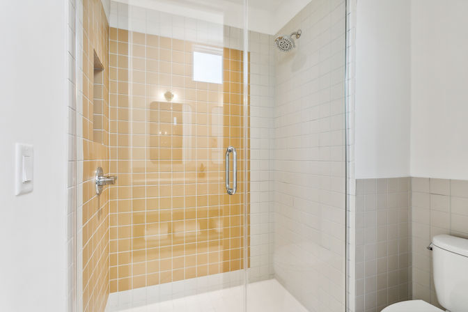 Sleek modern walk-in shower with glass door, beige and white square tiles, chrome rain showerhead and built-in niche, white toilet visible at the side