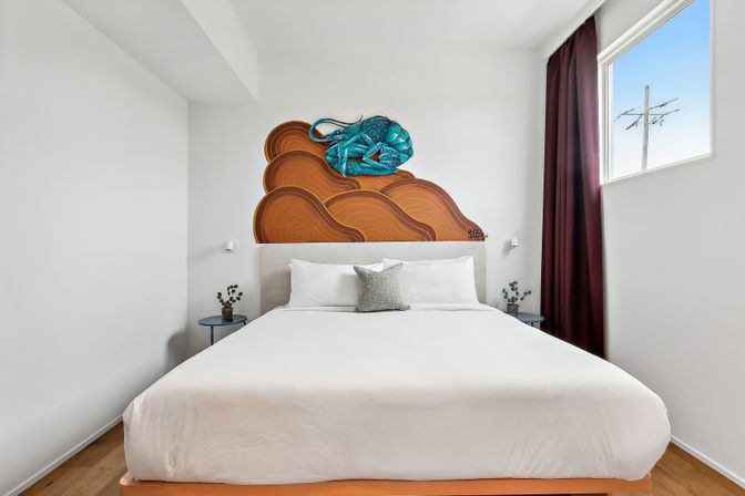 Bright minimalist bedroom with a large bed and white linens, gray accent pillow, teal sea-creature mural on orange wave shapes above the headboard, blue round nightstands with small potted plants, hardwood floor and a window with burgundy curtain letting in natural light.