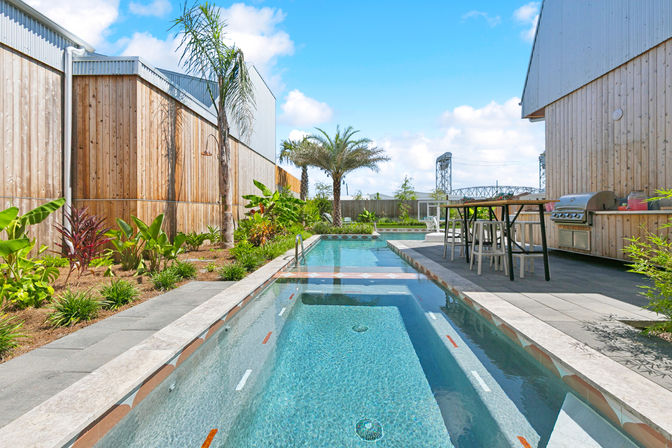 Sunny modern backyard lap pool with built-in spa, tropical palms and plants, gray paver patio, outdoor bar table with stools and stainless steel grill beside wood-clad fences.