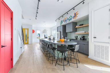 Bright sunlit modern kitchen with a long gray island seating a row of black barstools, mint-tile backsplash, gray cabinets, red interior door, yellow entry door and light hardwood floors.