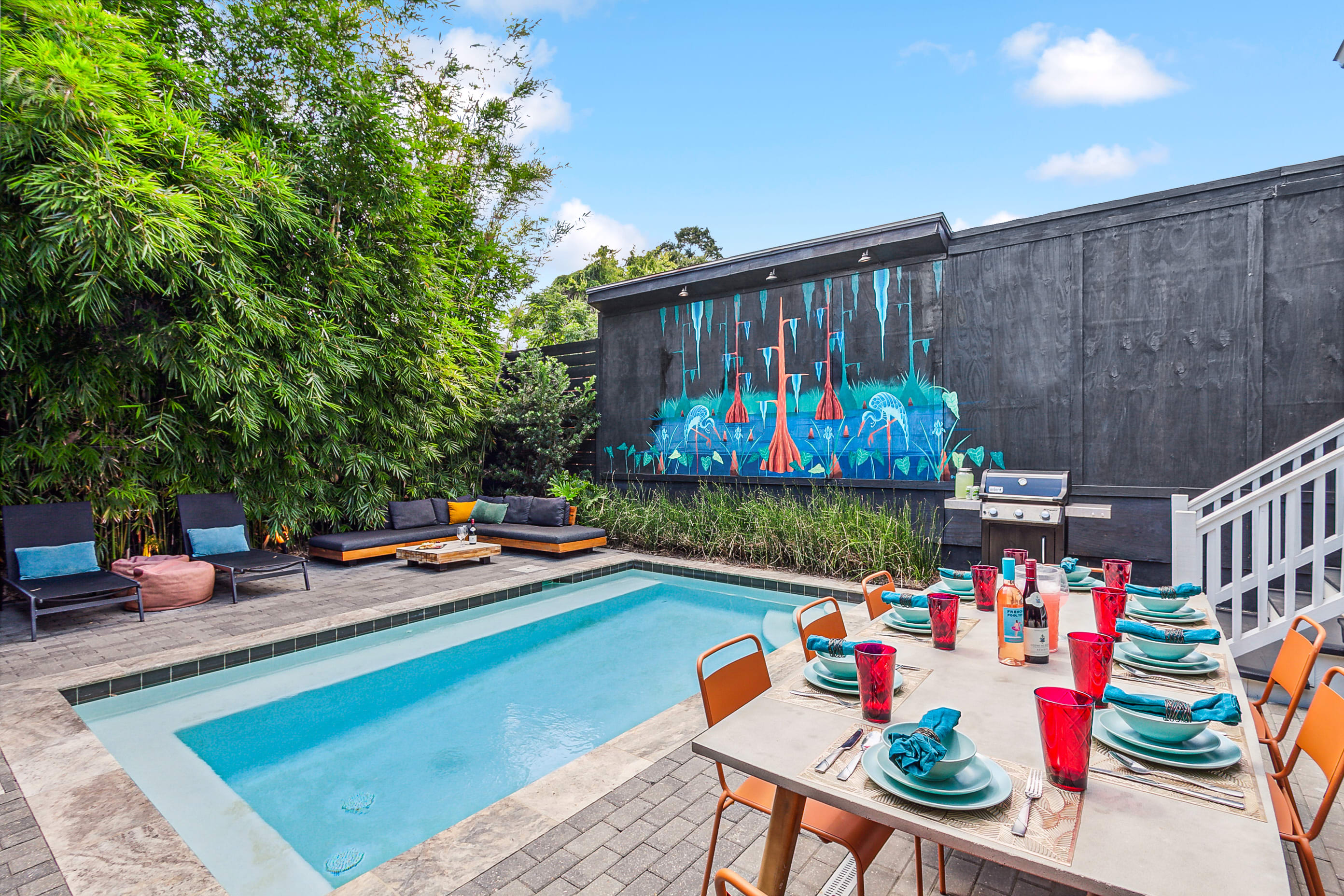 Modern backyard patio with small rectangular pool, colorful outdoor dining set and orange chairs, lounge seating by bamboo privacy, black wall with blue-orange mural and a grill