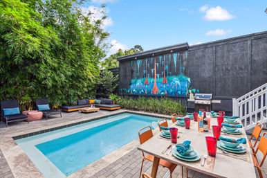 Modern backyard patio with small rectangular pool, colorful outdoor dining set and orange chairs, lounge seating by bamboo privacy, black wall with blue-orange mural and a grill