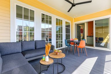 Bright covered porch with yellow siding, dark outdoor sectional sofa, round wooden side tables, colorful dining chairs around a table, ceiling fan and sliding glass doors.