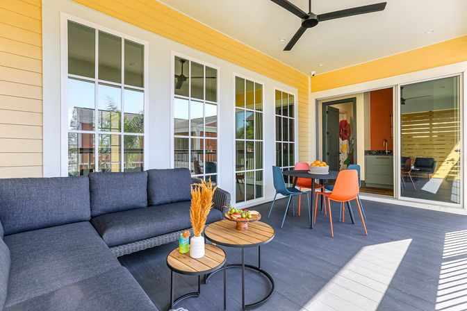 Bright covered porch with yellow siding, dark outdoor sectional sofa, round wooden side tables, colorful dining chairs around a table, ceiling fan and sliding glass doors.
