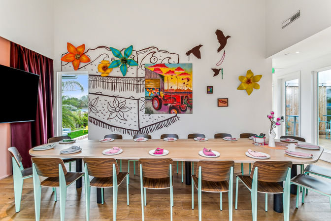 Bright, playful dining room with a long wooden table set for a meal, mint-accent chairs, colorful mural and oversized flower sculptures on a white wall, and large windows showing greenery.