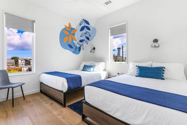 Bright modern guest room with two beds in white linens and blue throws, abstract blue-and-orange leaf wall mural, wood floors, minimalist chair and wall sconces, two windows showing an urban skyline and cloudy sky.