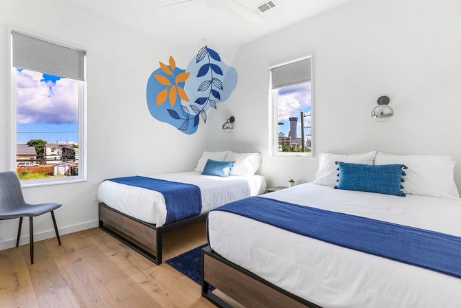 Bright modern guest room with two beds in white linens and blue throws, abstract blue-and-orange leaf wall mural, wood floors, minimalist chair and wall sconces, two windows showing an urban skyline and cloudy sky.