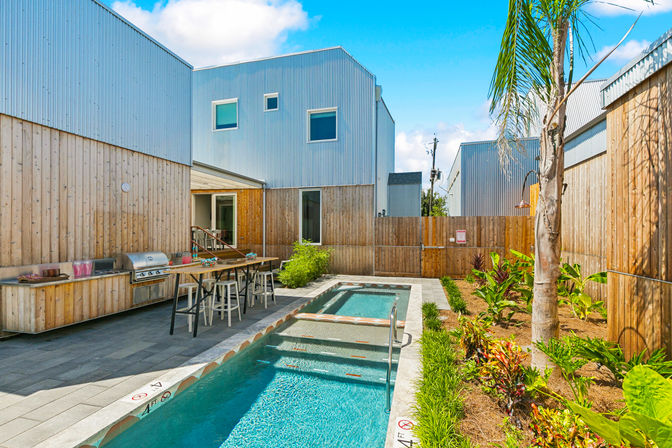 Sunlit modern courtyard with narrow lap pool, outdoor kitchen and bar seating, corrugated metal-clad townhouses, wooden fence and tropical landscaping