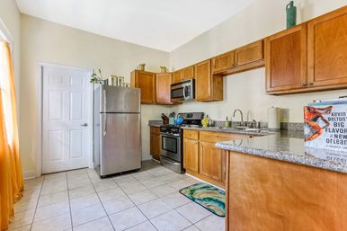 Cheerful sunlit kitchen with stainless-steel refrigerator and range, warm wood cabinets, granite countertops, tiled floor, colorful rug and orange sheer curtains