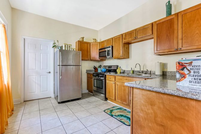 Cheerful sunlit kitchen with stainless-steel refrigerator and range, warm wood cabinets, granite countertops, tiled floor, colorful rug and orange sheer curtains