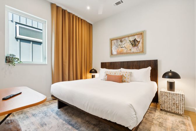 Stylish modern bedroom with king bed and crisp white linens, wood slatted headboard, decorative pillows, mustard floor-to-ceiling curtains, abstract wall art, black dome bedside lamps on textured cube nightstands, area rug and small window.