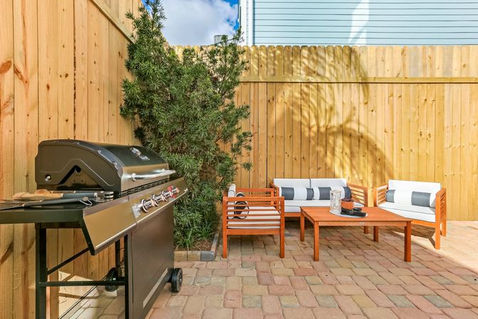 Sunny backyard patio with a black gas grill next to a wooden privacy fence, paver flooring, and a modern teak outdoor sofa set with white cushions and a coffee table, palm-frond shadow cast on the fence — ready for a relaxed cookout.