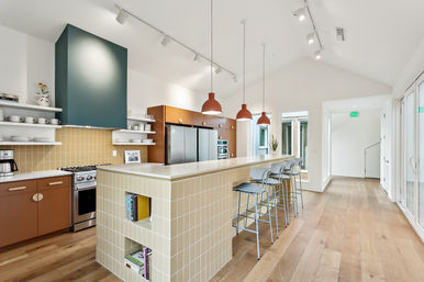 Bright open-plan modern kitchen with long tiled breakfast bar, three terracotta pendant lights, metal bar stools, stainless-steel appliances, open shelving and wide-plank wood floors.