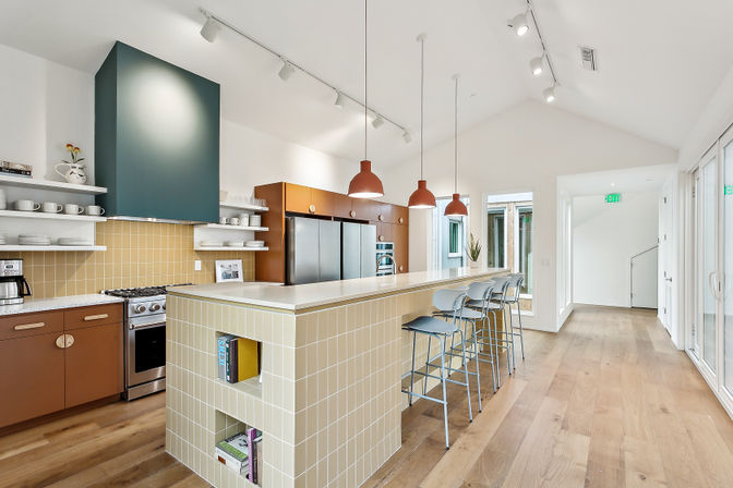 Bright open-plan modern kitchen with long tiled breakfast bar, three terracotta pendant lights, metal bar stools, stainless-steel appliances, open shelving and wide-plank wood floors.