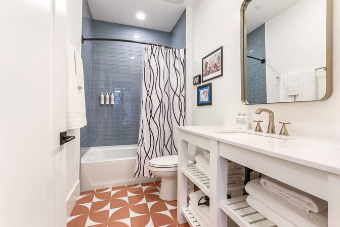Bright modern bathroom — white vanity with marble top and brass faucet, bathtub with blue subway-tile surround and wavy shower curtain, terracotta-and-white geometric floor tiles and open shelves of towels.