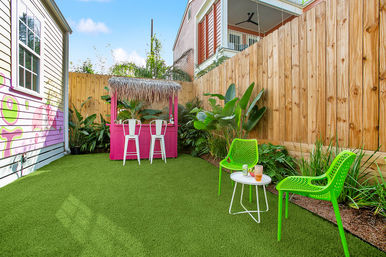 Colorful small backyard patio with a bright pink tiki bar and white bar stools, neon green chairs and a round table on green artificial turf, tropical plants and wooden privacy fence.