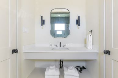 Minimalist modern bathroom vanity with white quartz countertop and undermount sink, matte black faucet and hardware, rounded rectangular mirror flanked by wall sconces, neatly folded white towels and a hair dryer on the open shelf below.