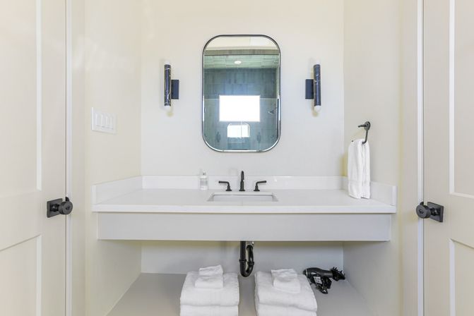 Minimalist modern bathroom vanity with white quartz countertop and undermount sink, matte black faucet and hardware, rounded rectangular mirror flanked by wall sconces, neatly folded white towels and a hair dryer on the open shelf below.