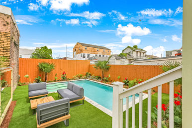 Cozy urban backyard with rectangular turquoise swimming pool, artificial turf lawn, gray modern patio lounge set, wooden privacy fence and tropical plants under a bright blue sky.