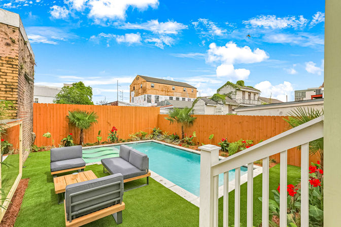 Cozy urban backyard with rectangular turquoise swimming pool, artificial turf lawn, gray modern patio lounge set, wooden privacy fence and tropical plants under a bright blue sky.