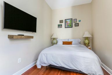 Cozy small bedroom with white bedding and orange accent pillow, wall-mounted flat-screen TV, hardwood floors, framed gallery wall and matching bedside lamps.