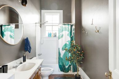 Small modern gray bathroom with white tub and toilet, tropical green-leaf shower curtain, round mirror above wooden vanity, cheery potted citrus plant and brass wall hooks