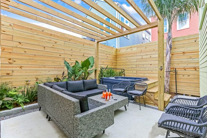 Cozy urban backyard patio with wooden pergola and horizontal privacy fence, gray wicker sectional, hot tub, black outdoor chairs and tropical plants.