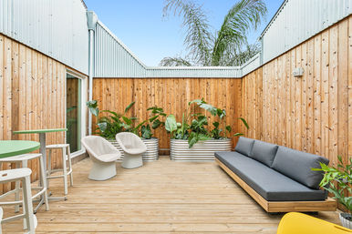 Sunny urban patio deck with cedar fencing and corrugated metal walls, modern gray sofa, two white lounge chairs, green bistro table, and galvanized planters filled with tropical plants and a palm peeking above.