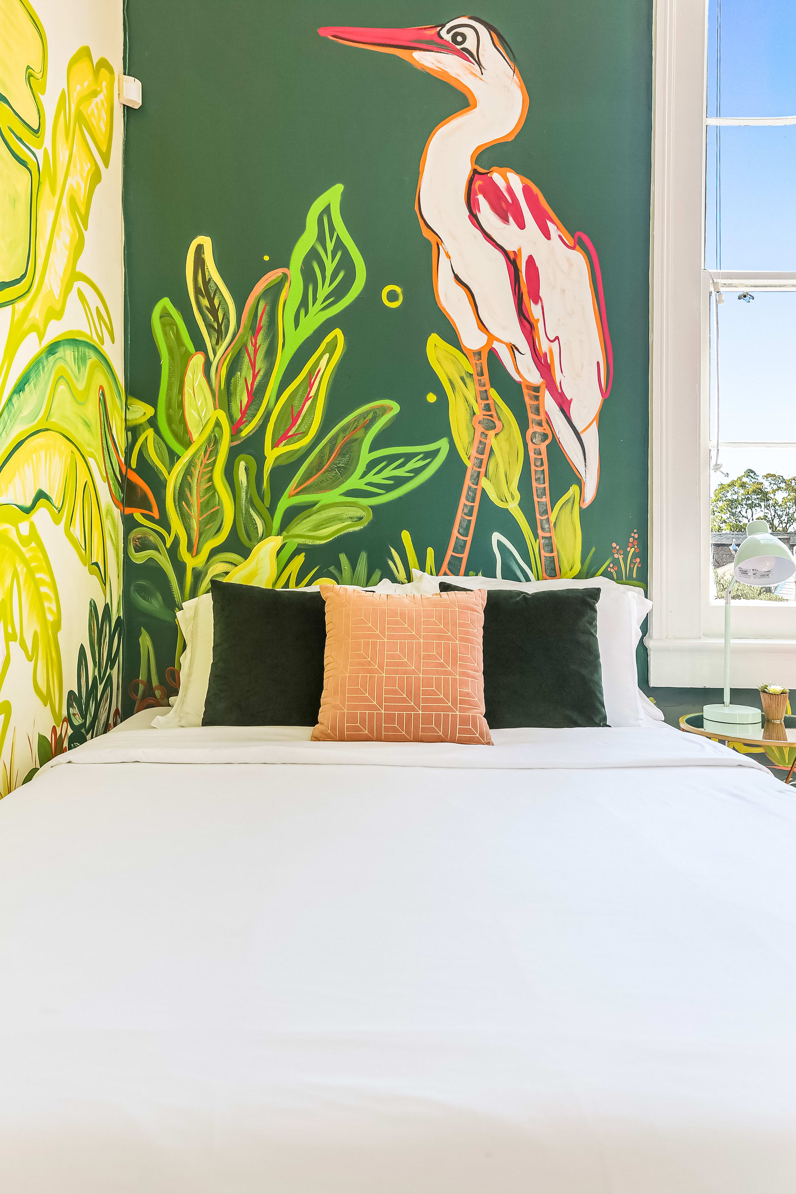 Sunlit bedroom with white bedding, dark green pillows and a peach accent cushion in front of a bold tropical heron and leafy wall mural beside a window