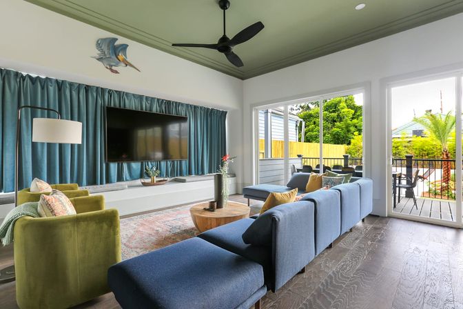 Bright modern living room with blue sectional and green armchairs, wall-mounted TV, black ceiling fan, hardwood floors and large sliding glass doors opening to a sunny balcony and landscaped yard