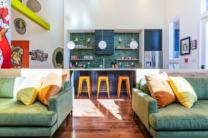Bright modern open-concept living room and kitchen with high ceilings, green velvet sofas with colorful pillows, three yellow bar stools at a dark island, glossy green tile backsplash and round pendant lights.
