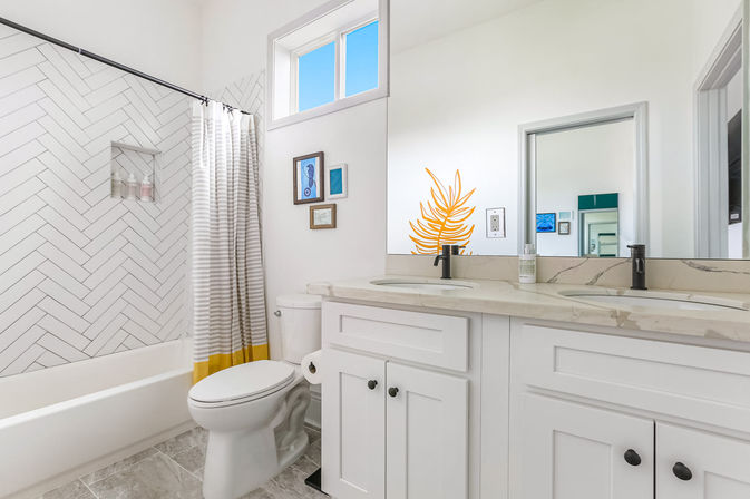 Bright modern white bathroom with marble countertop and double sinks, black faucets, herringbone-tiled bathtub-shower, gray-and-yellow striped curtain, toilet, and framed wall art.