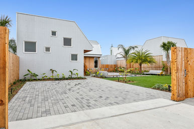 Contemporary white corrugated metal house with angled roof opening to a fenced courtyard — gray paver driveway, manicured lawn, palm trees and outdoor lounge seating under a clear blue sky.