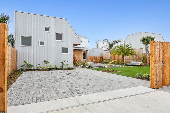 Contemporary white corrugated metal house with angled roof opening to a fenced courtyard — gray paver driveway, manicured lawn, palm trees and outdoor lounge seating under a clear blue sky.