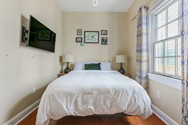 Cozy sunlit bedroom with crisp white bedding on a centered bed, wall-mounted TV, framed art above the headboard, matching bedside lamps, patterned curtains and warm hardwood floors.