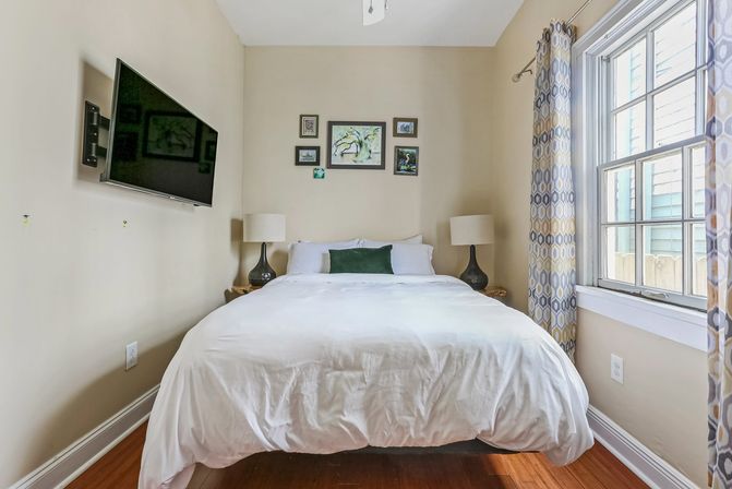 Cozy sunlit bedroom with crisp white bedding on a centered bed, wall-mounted TV, framed art above the headboard, matching bedside lamps, patterned curtains and warm hardwood floors.