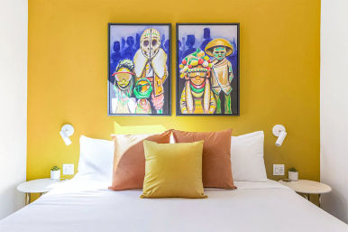 Bright modern bedroom with mustard-yellow accent wall, white bedding, mustard and terracotta throw pillows, two colorful tribal-inspired framed paintings above the bed, round white bedside tables with succulents and wall sconces.