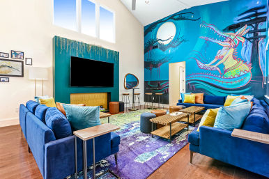 Vibrant modern living room with high ceilings and large windows, teal media wall and whimsical dragon mural, blue velvet sectional sofas with colorful throw pillows, wooden coffee tables and a purple-green area rug.