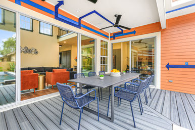 Modern covered backyard patio outdoor dining area with gray table and navy woven chairs on gray deck, orange siding with blue arrow mural, ceiling fan, and sliding glass doors into the living room