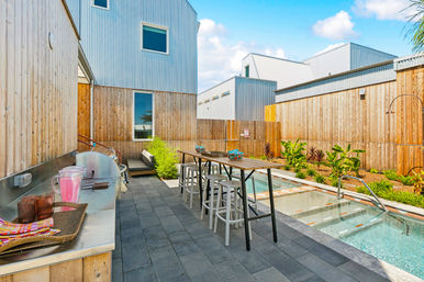 Sunny modern backyard patio with narrow lap pool, tall wooden fences and corrugated metal siding, long high bar table with stools, outdoor grill counter, lounge seating and tropical plants on paver decking.