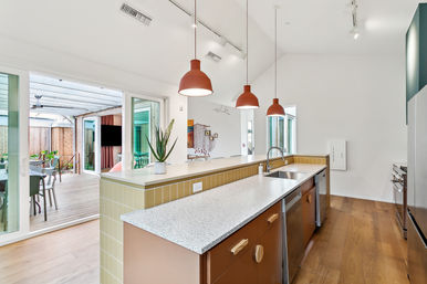 Bright modern open-plan kitchen with a long speckled island, stainless sink, three terracotta pendant lights, honey wood floors, and sliding glass doors opening to a covered wooden deck patio for indoor-outdoor living.