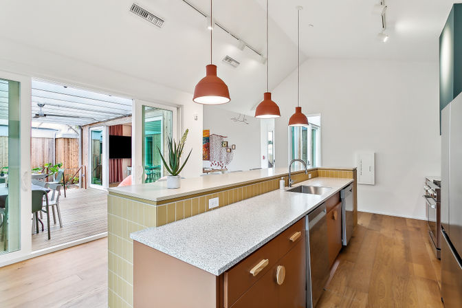 Bright modern open-plan kitchen with a long speckled island, stainless sink, three terracotta pendant lights, honey wood floors, and sliding glass doors opening to a covered wooden deck patio for indoor-outdoor living.